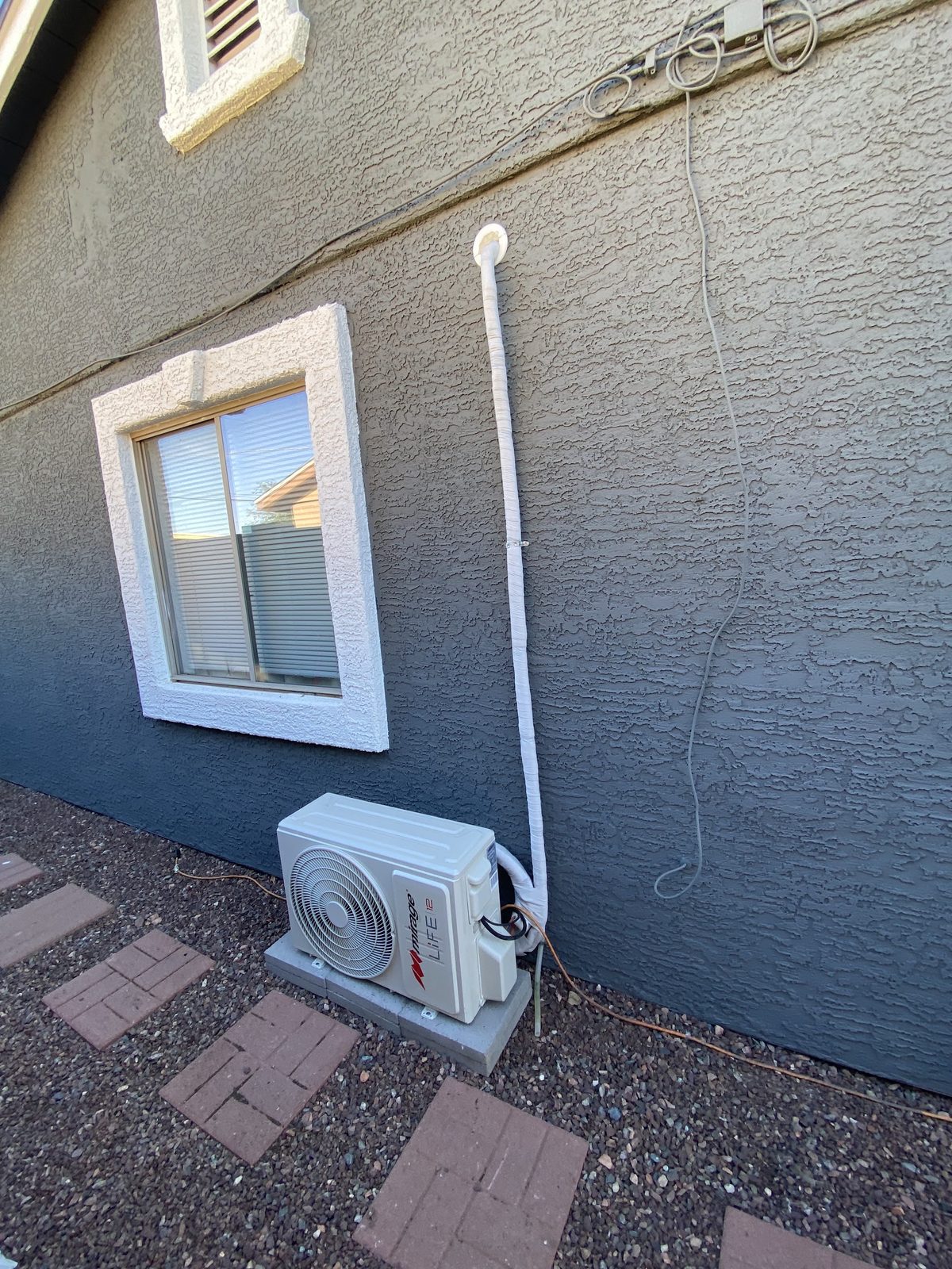 A mini split condenser unit set on pads against a two tone stucco wall, with the line set wrapped and run neatly up to the roof line.