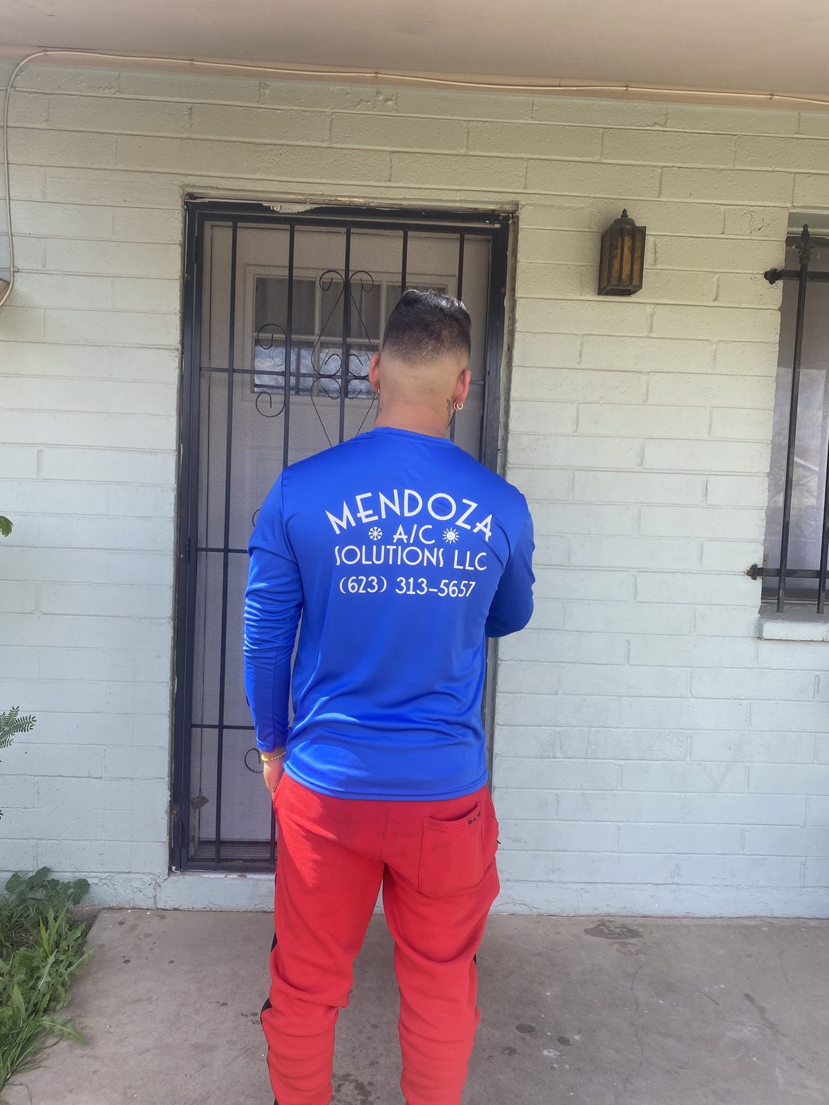 A Mendoza A/C Solutions technician at a customer's front door, the company name and phone number printed across the back of his blue work shirt.